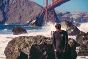 man standing beside black stone with sea waves near red metal bridge during daytime